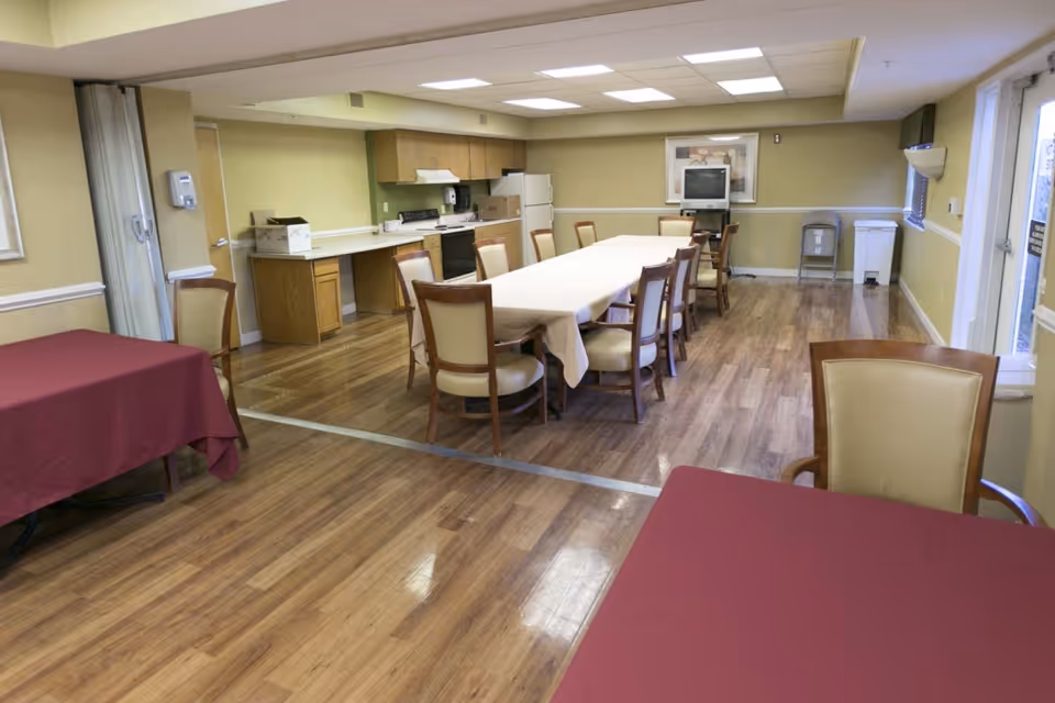 Interior dining/activity room with a long table and chairs, a small kitchen area in the back, and wood flooring.