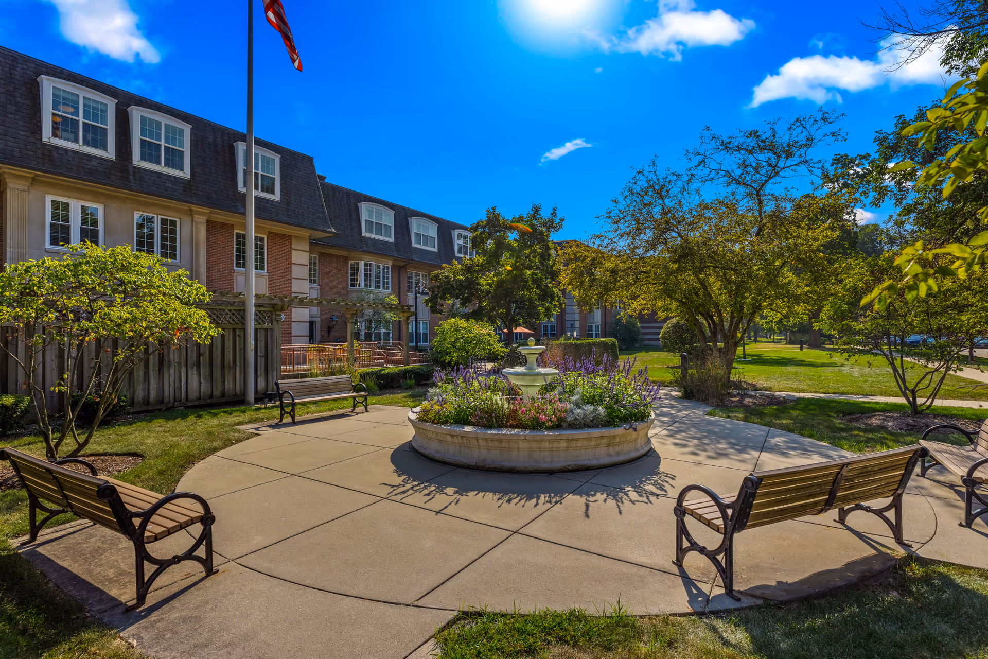 Outdoor courtyard area at Three Crowns Park featuring a circular flower bed with a small fountain in the center, surrounded by benches and trees. The building with multiple windows and an American flag on a flagpole are visible in the background under a bright blue sky with a few clouds.