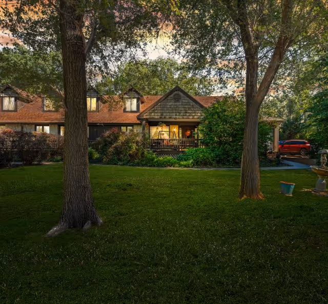 A charming countryside home with a porch surrounded by lush greenery and trees, viewed from a grassy lawn during sunset. A red car is parked on the driveway to the right.