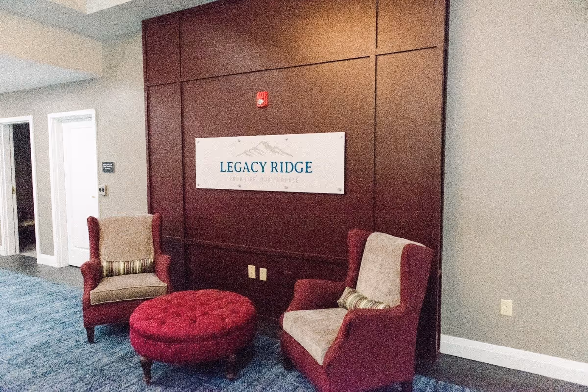 Small lounge area with two upholstered armchairs, a round ottoman, and a 'Legacy Ridge' sign on a dark wood-paneled wall.