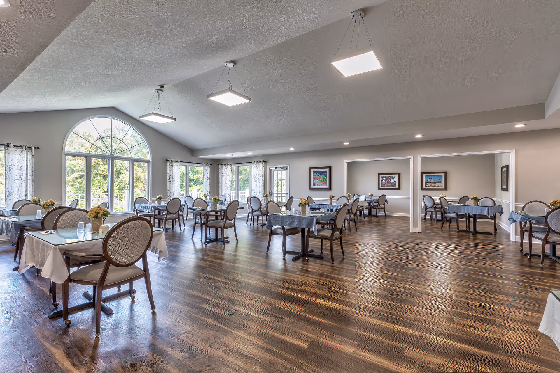 Spacious dining room with multiple tables covered with tablecloths and surrounded by chairs. Large arched window and several smaller windows allow natural light to fill the room. The floor is wooden, and the walls are decorated with framed artwork. Ceiling lights provide additional illumination.