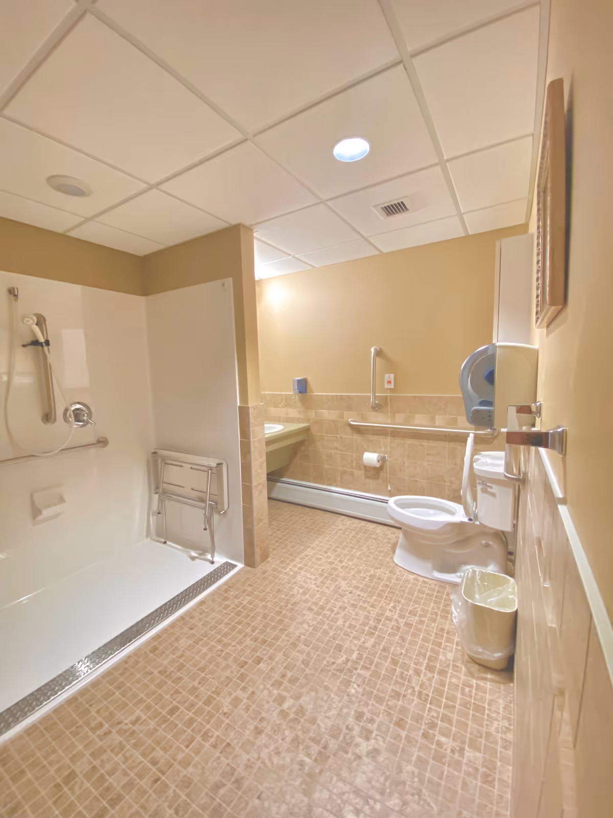 A spacious bathroom with beige tiled floor and walls. The room features a walk-in shower with a foldable seat and grab bars, a toilet with grab bars, a wall-mounted paper towel dispenser, a small trash can, and a sink with a mirror above it. The ceiling has recessed lighting and ventilation.