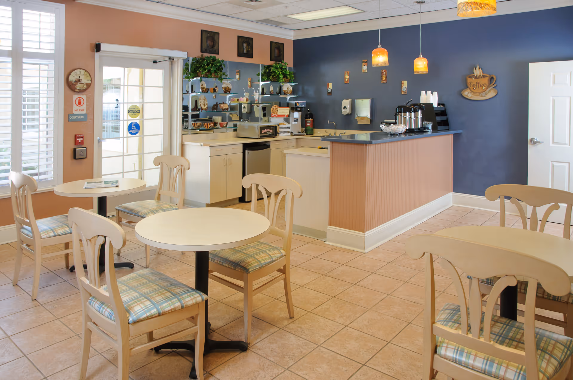 A small dining area with round tables and wooden chairs with plaid cushions. The room features a coffee bar with coffee dispensers, cups, and a sink. The walls are painted in peach and blue tones, decorated with small wall hangings and plants on shelves behind the counter. There is a door and large windows with white shutters letting in natural light.