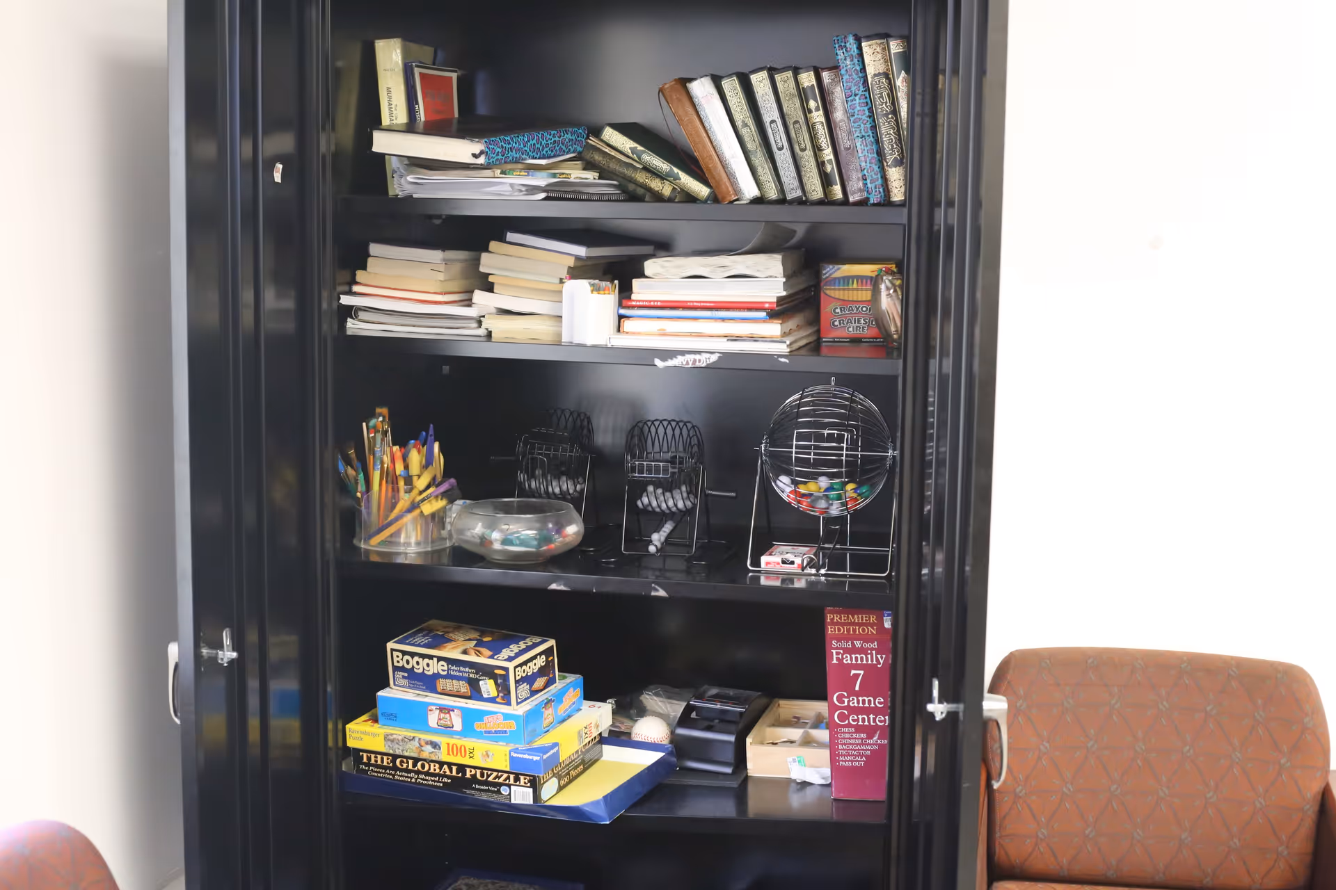 A black cabinet filled with various books, notebooks, board games including Boggle and The Global Puzzle, bingo cages with balls, a container with colored pencils, and other small items. To the right of the cabinet is a brown cushioned chair with a patterned fabric.