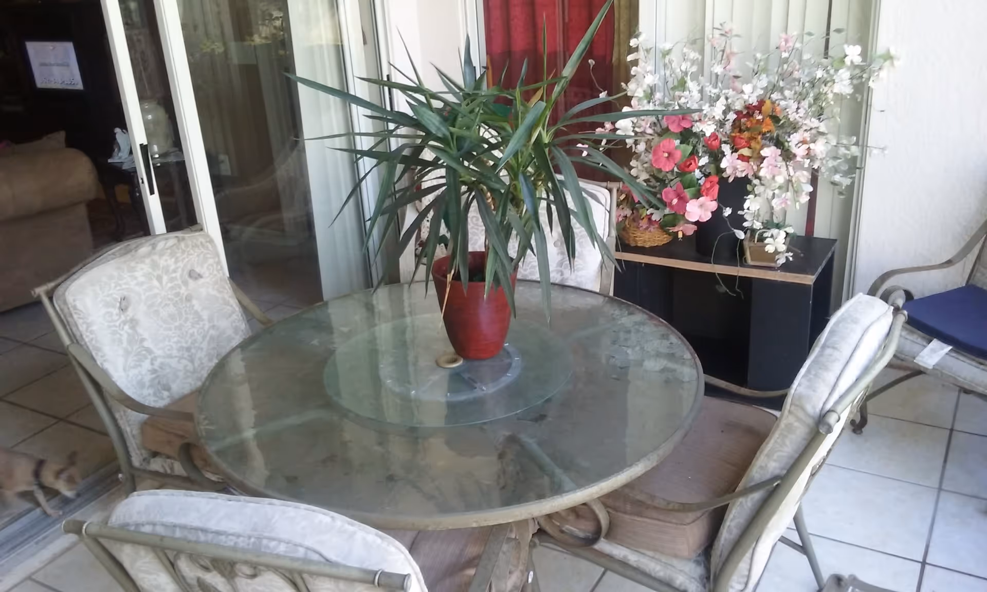 A round glass-top table with a red potted plant in the center, surrounded by four cushioned chairs. Behind the table is a black shelf with two flower arrangements, one with pink and white flowers and the other with mixed colors. The setting appears to be an indoor or enclosed patio area with tiled flooring and sliding glass doors leading to another room with a couch visible.