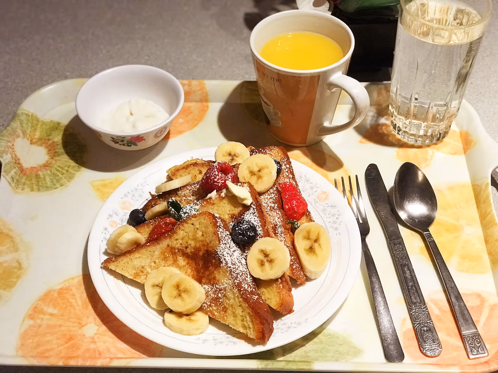 A tray with French toast topped with banana slices and berries, a mug of orange juice, a glass of water, a small bowl and utensils.