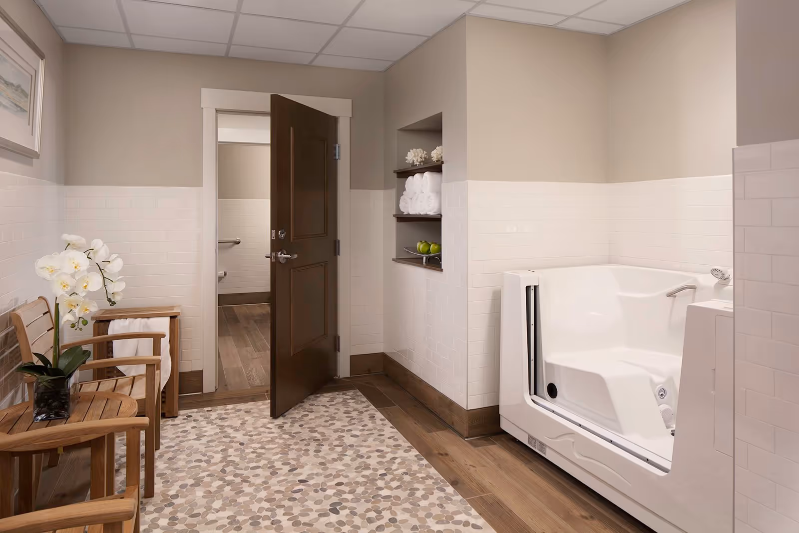 A clean and modern bathroom featuring a walk-in bathtub with a door, a wooden chair with a white towel draped over it, a small wooden table with a white orchid plant, built-in shelves holding rolled white towels and green apples, and a partially open dark wooden door leading to another room with a grab bar visible.