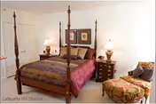 A well-lit bedroom featuring a four-poster bed with a patterned bedspread and multiple pillows. On either side of the bed are wooden nightstands with lamps and decorative items. A cushioned armchair with a matching ottoman is positioned near the window, and framed artwork hangs on the wall above the bed.