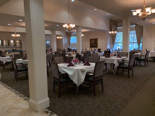A spacious dining room with multiple tables covered in white tablecloths, each set with folded purple napkins, glassware, and floral centerpieces. The room features carpeted floors, large windows with curtains, and multiple chandeliers providing warm lighting.