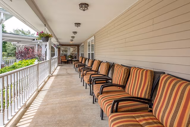 A long covered porch with a row of striped cushioned chairs along one side and white railing on the other, overlooking greenery and trees.