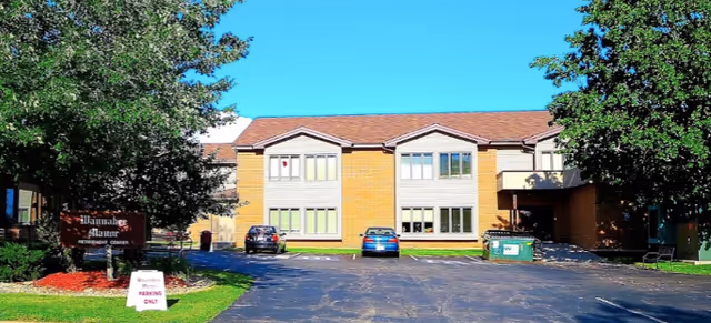 Front exterior view of Waunakee Manor Health Care Center building with a parking lot in front, two cars parked, trees on both sides, and a sign indicating parking only.