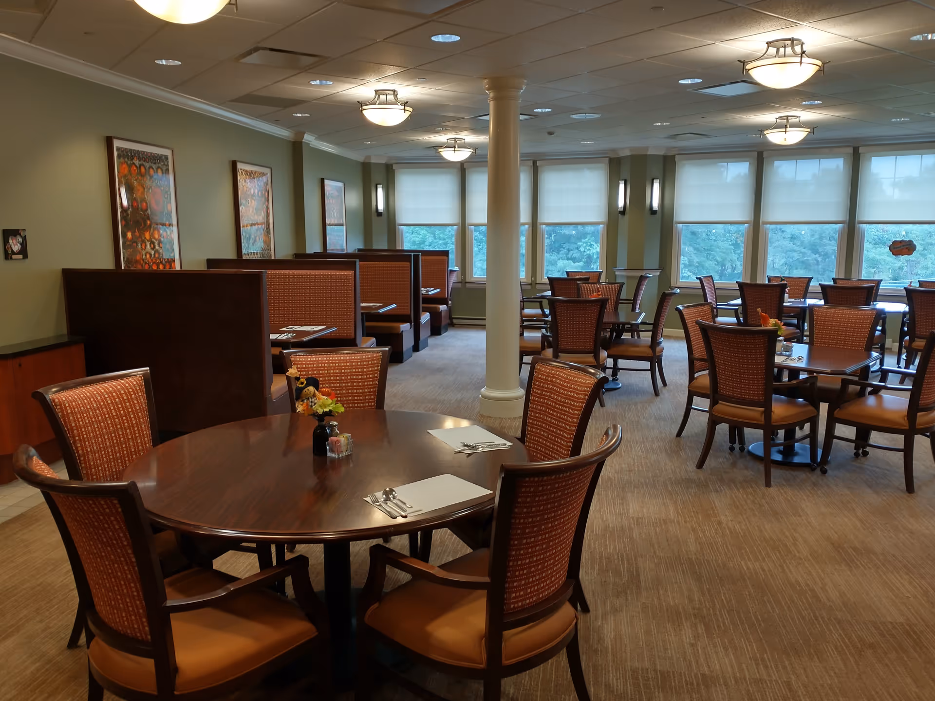 A spacious dining room with multiple wooden tables and chairs arranged neatly. The room has large windows with white blinds allowing natural light to enter. There are booth seating areas along one wall with framed artwork above them. The ceiling has recessed lighting and hanging light fixtures.