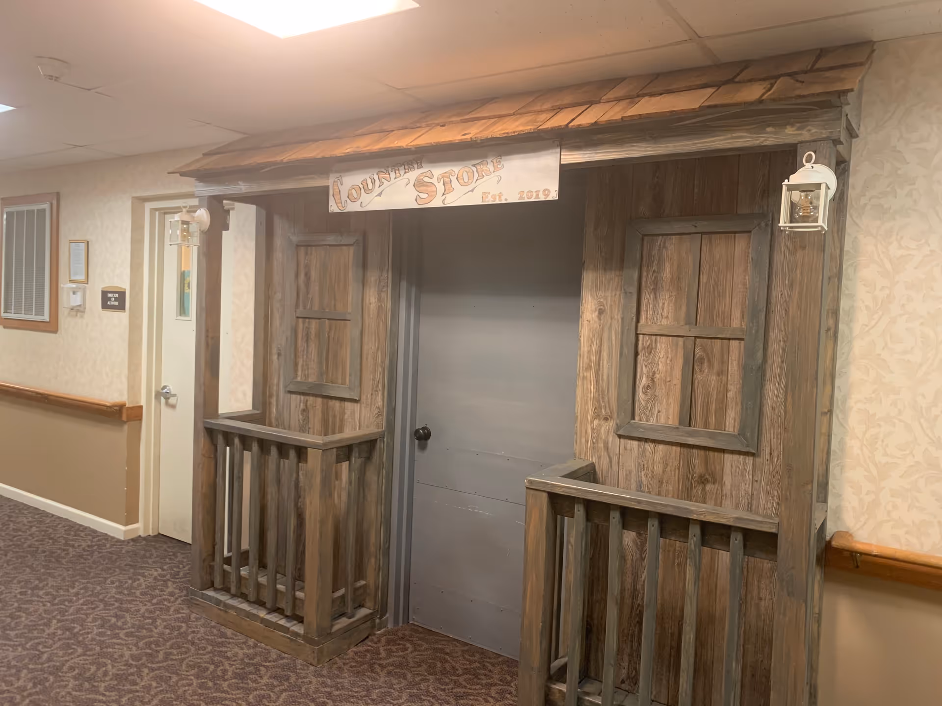 Indoor hallway with a rustic wooden structure resembling a small storefront with a sign that reads 'Country Store Est. 2019'. The structure has two window frames and small railings on either side of a closed gray door. The hallway has beige patterned wallpaper and carpeted floor.