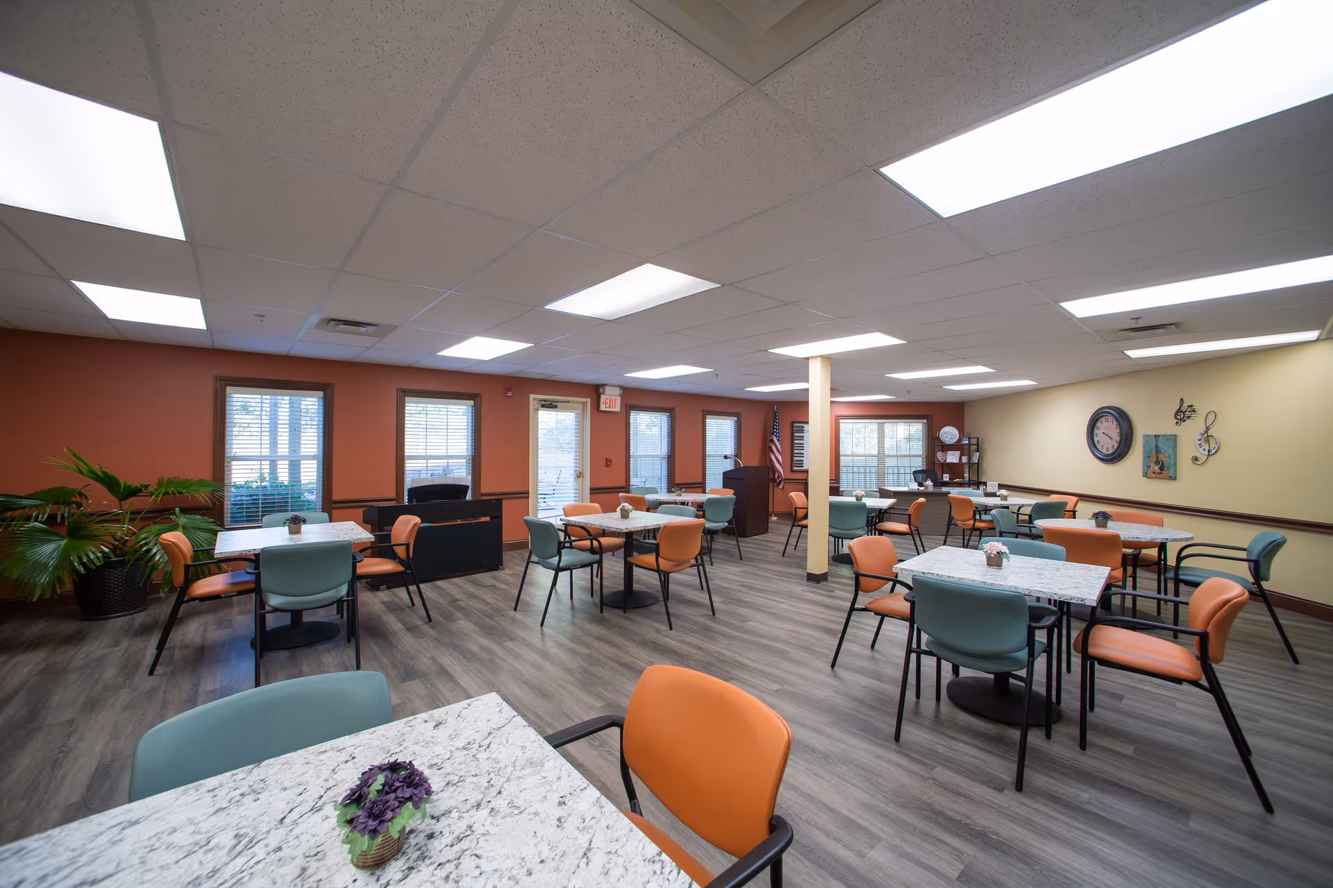 Communal dining room with marble-top tables, orange and teal chairs, plants, and wall decor.