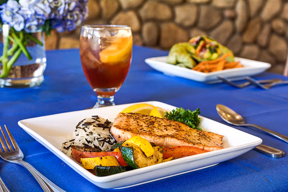 A plated meal on a white square plate featuring grilled salmon, wild rice, and roasted vegetables including zucchini and yellow squash, garnished with lemon wedges and parsley. In the background, there is a glass of iced tea with a lemon slice and another plate with a wrap and sweet potato fries on a blue tablecloth with a stone wall backdrop.