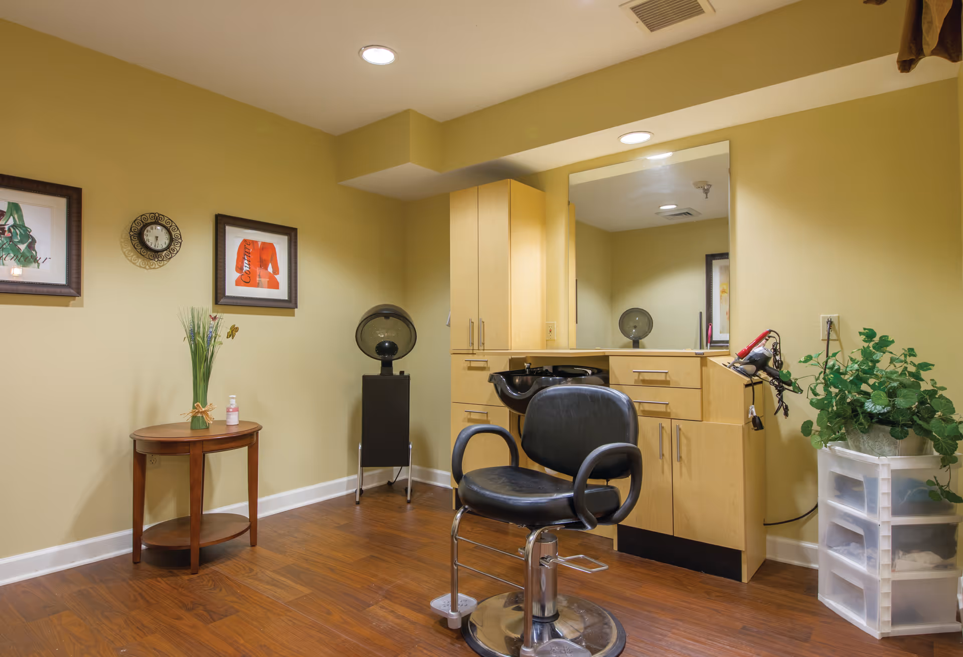 Small on-site salon area with a styling chair, shampoo basin, mirror, cabinetry and a side table against yellow walls.
