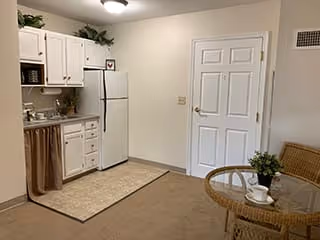 Small kitchen area with white cabinets, a white refrigerator, microwave, and a sink. There is a beige rug on the floor in front of the kitchen area. To the right, there is a round glass table with two wicker chairs, one of which has a small potted plant and a white cup and saucer on the table. A closed white door is visible on the wall behind the table.
