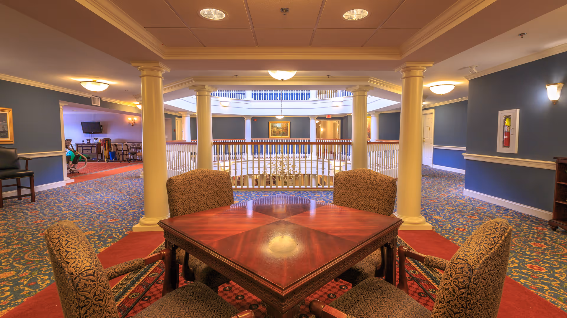 Interior view of a senior living facility featuring a square wooden table with four upholstered chairs arranged around it. The area has patterned carpet flooring and is surrounded by white columns and blue walls. In the background, there is a railing overlooking a lower level with a chandelier, and additional seating and decor are visible.