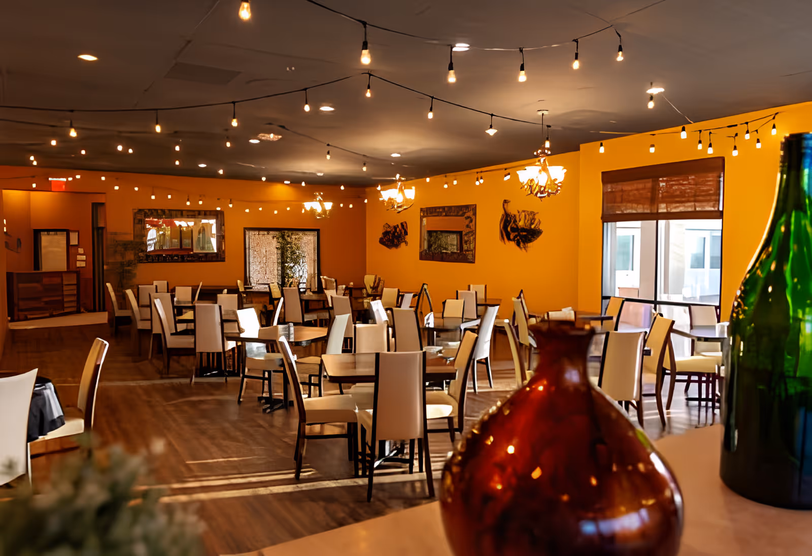 Spacious, warmly lit dining room with multiple tables and chairs, string lights overhead, and decorative bottles in the foreground.