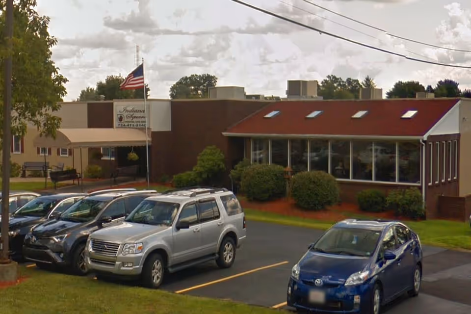 Exterior view of Heritage Grove at Indiana facility showing a single-story brick building with a red roof section and several windows. There is an American flag on a flagpole near the entrance, and several cars are parked in the parking lot in front of the building. Trees and shrubs surround the area under a cloudy sky.