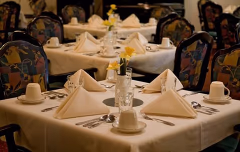 A dining room table set for a meal with white tablecloth, folded white napkins, cups, glasses, silverware, and small vases with yellow flowers. Several similar tables and chairs with patterned upholstery are visible in the background.