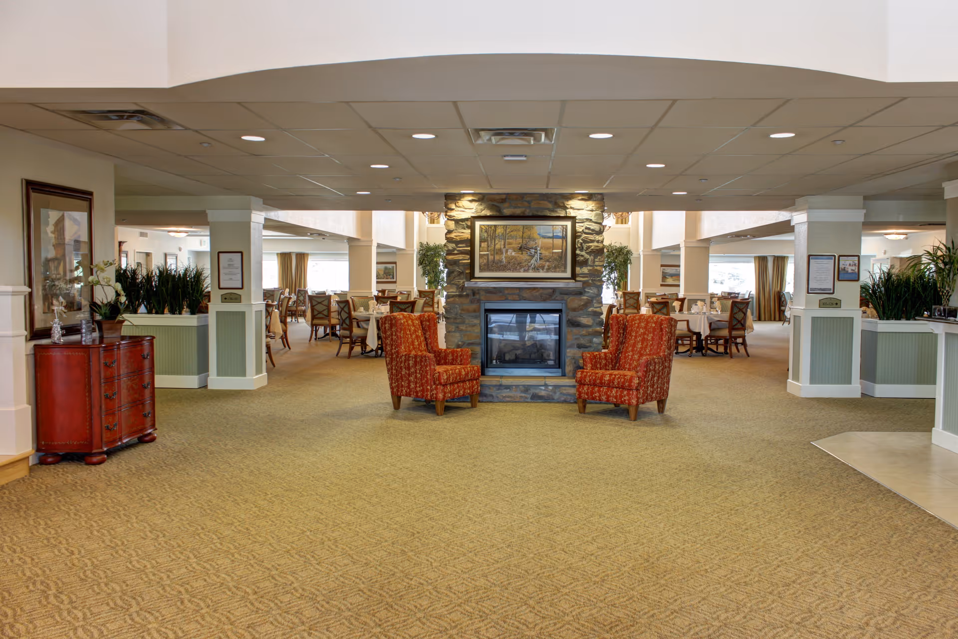 Interior view of a senior living facility dining area with a stone fireplace in the center, flanked by two red patterned armchairs. The room has carpeted floors, several dining tables with chairs, potted plants, and framed artwork on the walls.