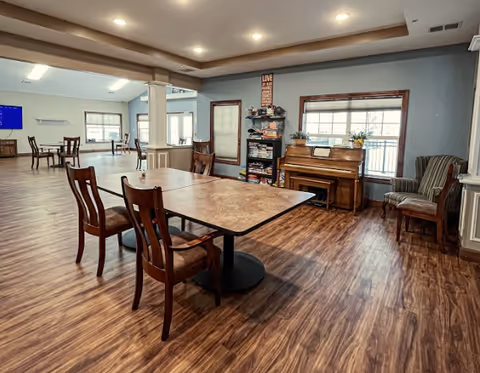Spacious communal dining/activity room with tables and chairs, a piano, bookshelf, and large windows.
