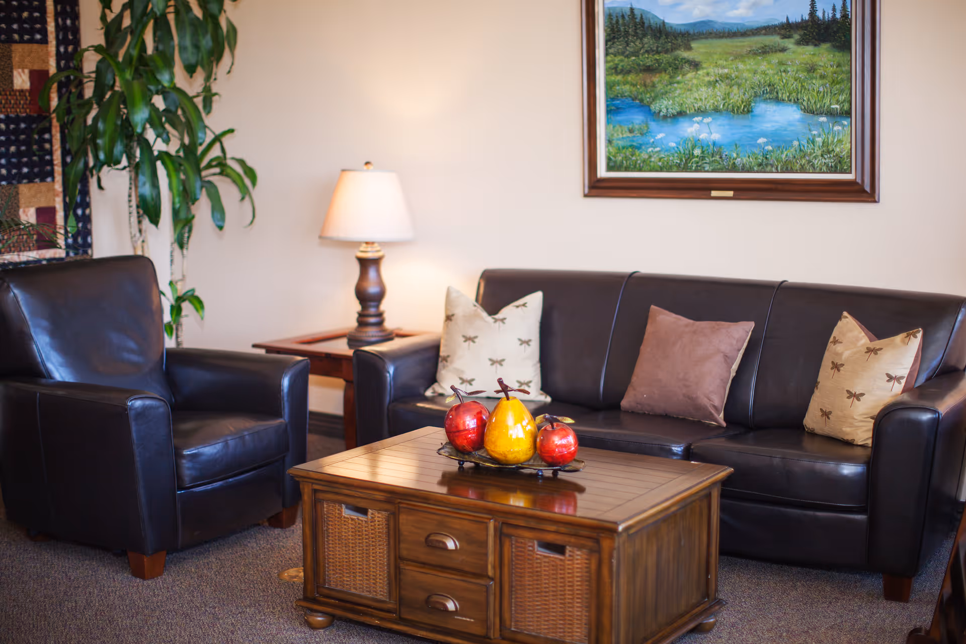 Cozy sitting area with a leather sofa and armchair, wooden coffee table topped with decorative fruit, a side lamp, and a framed landscape painting.