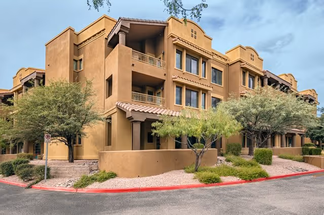 Exterior view of a multi-story senior living facility building with beige stucco walls, balconies, and a tiled roof. The building is surrounded by desert landscaping with small trees, bushes, and gravel. The sky is partly cloudy.