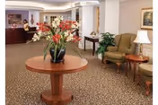 Reception lobby with a round table holding a floral arrangement, seating area with armchairs and a lamp, and a reception desk in the background.