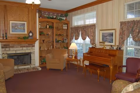 A cozy living room with a stone fireplace on the left, wooden built-in shelves filled with decorative items, two armchairs, a wooden piano with a matching bench, a side table with a lamp, and windows with floral curtains letting in natural light.