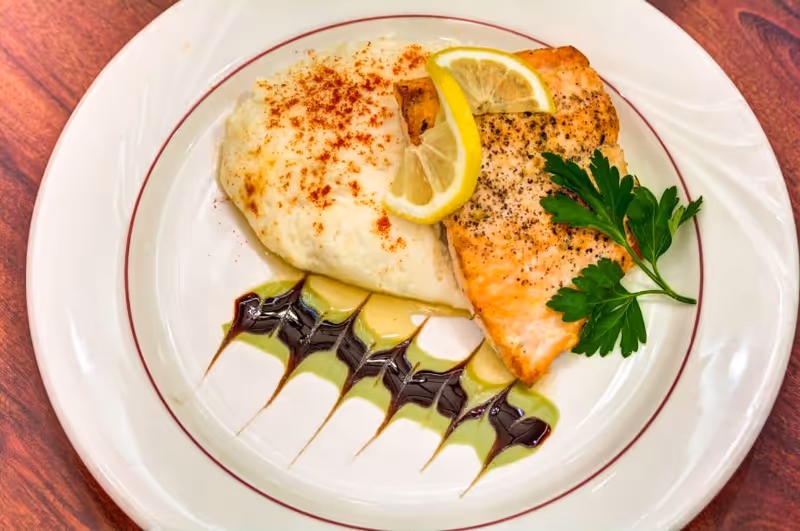 A plated meal featuring a piece of seasoned cooked salmon garnished with two lemon slices and a sprig of parsley, served alongside a portion of mashed potatoes sprinkled with paprika, with decorative drizzles of green and dark sauces on a white plate with a red rim, placed on a wooden table.