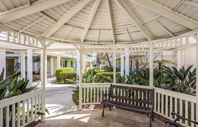 View from inside a white wooden gazebo with a bench, overlooking a garden area with green plants and bushes, surrounded by a senior living facility building.