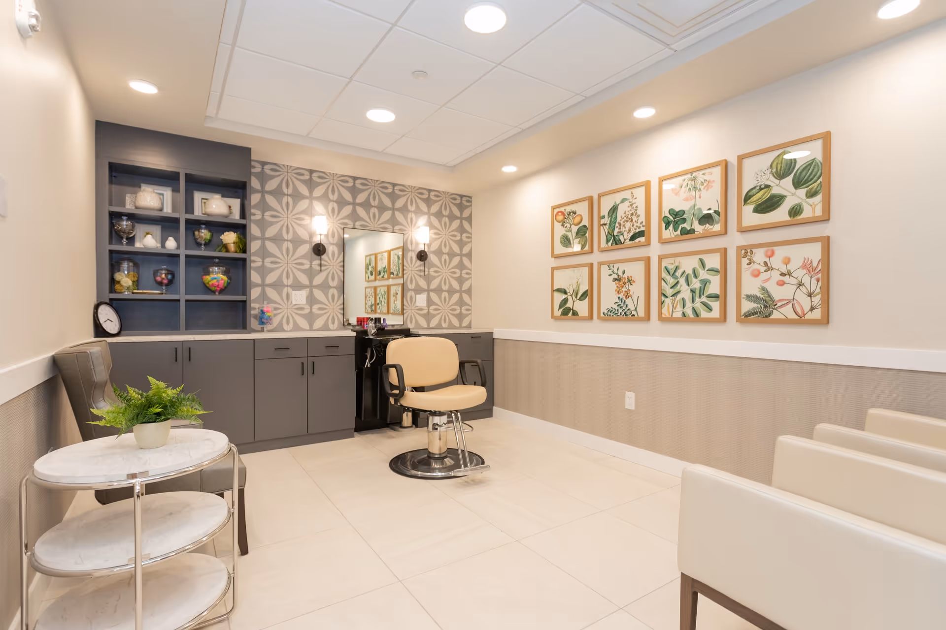 A bright and clean salon area in a senior living facility with a beige salon chair in front of a mirror mounted on a wall with patterned tiles. To the left, there is a dark gray cabinet with shelves holding decorative items and jars. On the right wall, there are eight framed botanical prints arranged in two rows. The floor is tiled in light beige, and there are several cream-colored chairs along the right side. A small round table with a potted plant is positioned near a gray upholstered chair on the left.