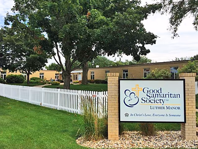 Exterior view of Good Samaritan Society - Luther Manor facility with a white picket fence, green lawn, trees, and a sign displaying the facility's name and slogan.