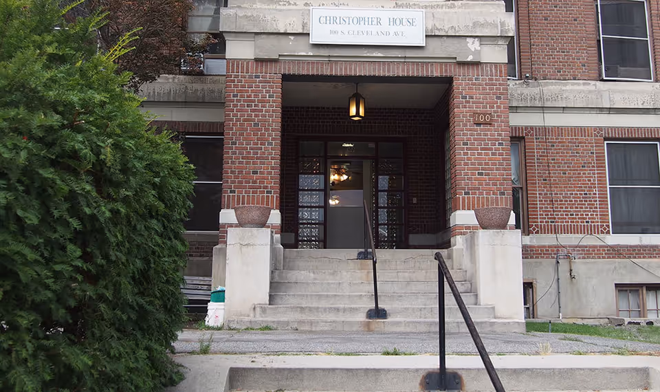 Front entrance of a brick building named Christopher House with stairs leading up to double glass doors, flanked by large planters and greenery on the left side.