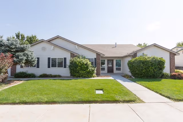 Front exterior view of a single-story senior living facility building with white siding, brick accents, black shutters, a well-maintained green lawn, bushes, and a concrete walkway leading to the entrance under a clear sky.