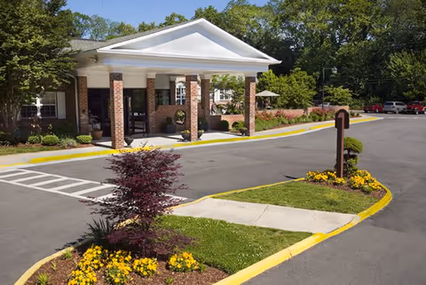 Front exterior view of a brick building with a white portico supported by columns, surrounded by landscaped greenery and flower beds, with a driveway and parking area in front.