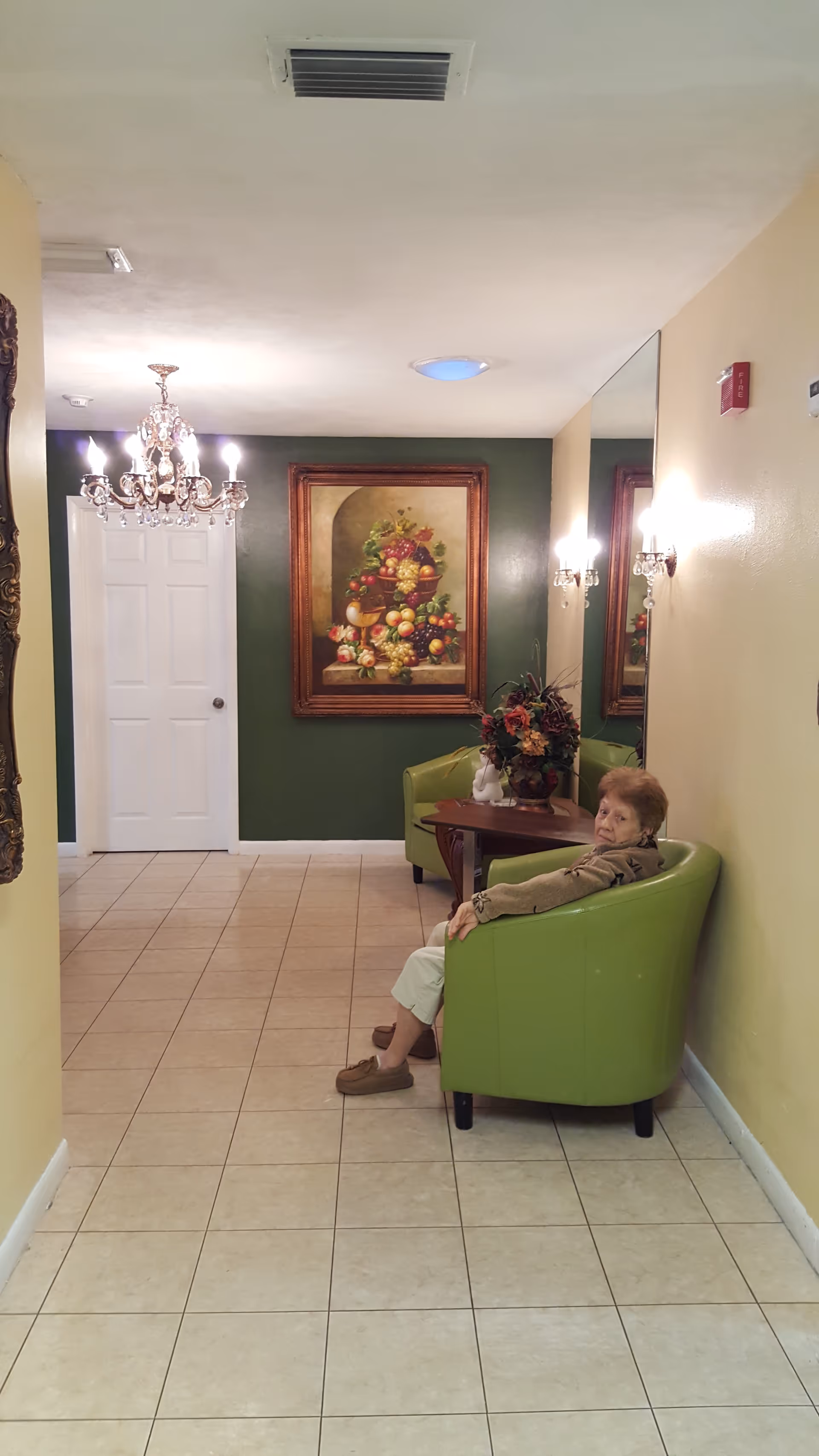 A hallway in Cameron Assisted Living Facility with tiled floor and light yellow walls. An elderly woman is sitting in a green armchair next to a small wooden table with a floral arrangement. The wall behind her is green and features a large framed painting of a fruit basket. A chandelier and wall sconces provide lighting, and a large mirror is mounted on the right wall.