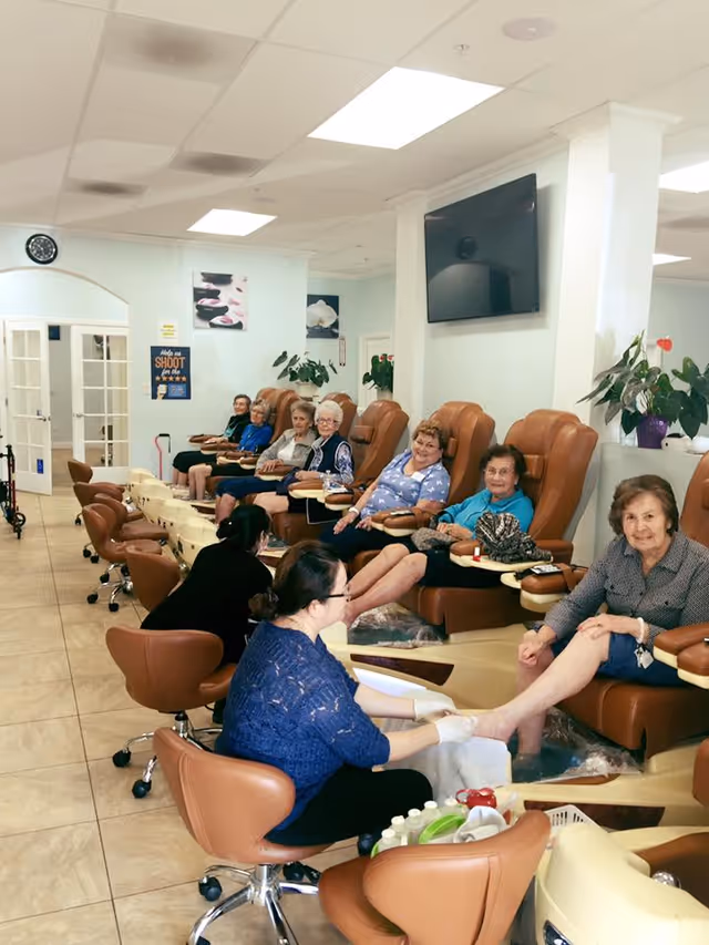 Several older women sit in a row of pedicure chairs while staff provide foot treatments in a bright salon-like room.