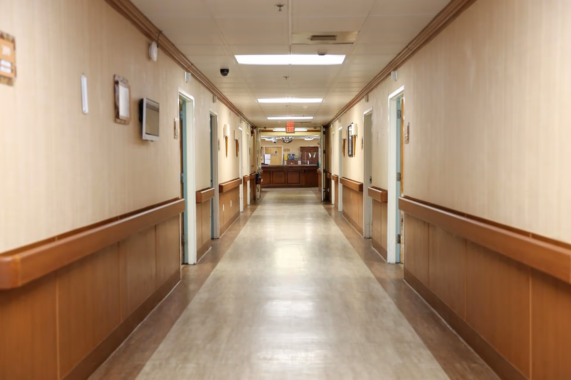 Long, well-lit corridor in a senior living facility with doors along both sides and a reception desk at the far end.