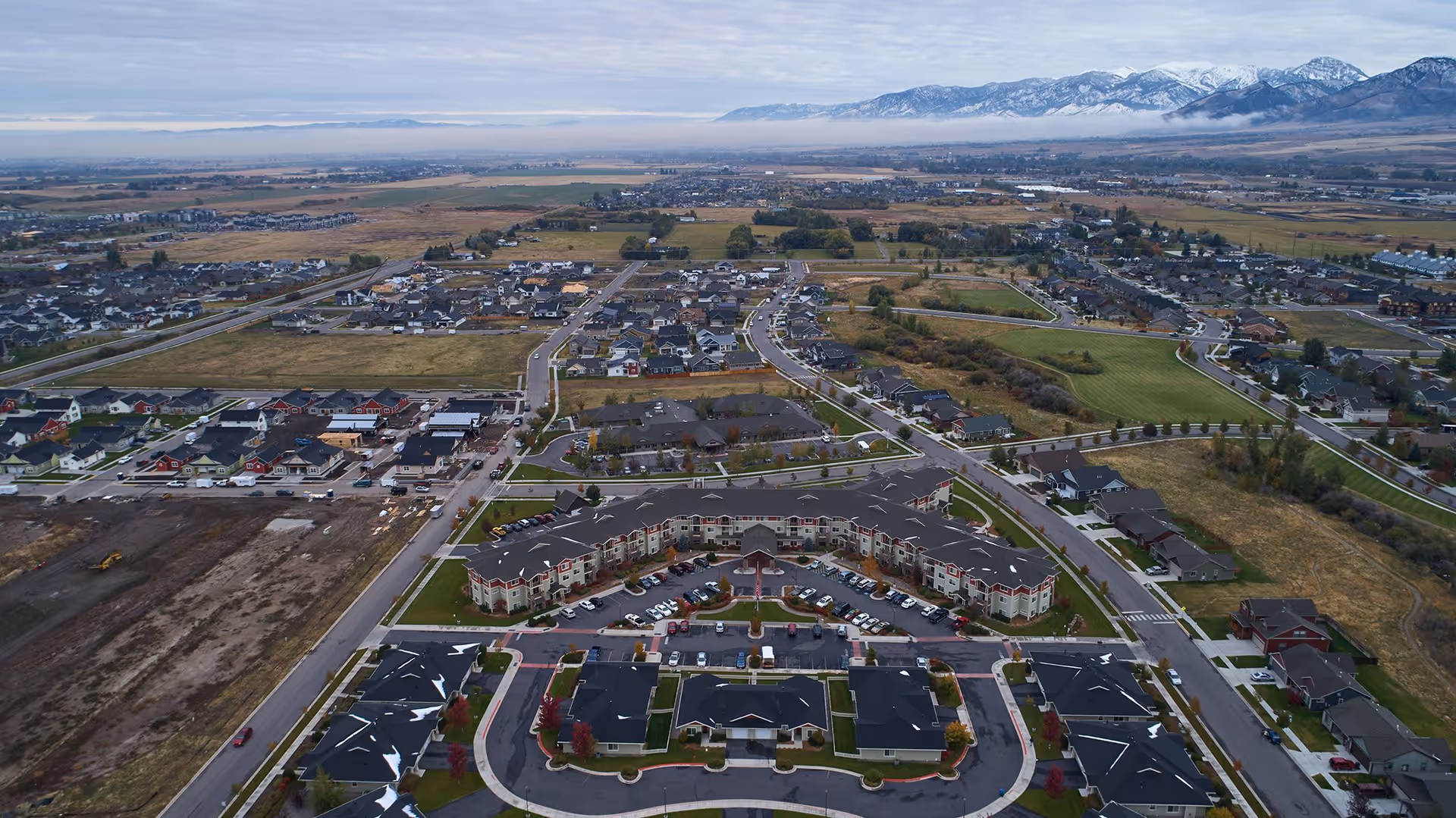 Aerial view of Bozeman Lodge facility surrounded by residential neighborhoods, open fields, and distant snow-capped mountains under a cloudy sky.