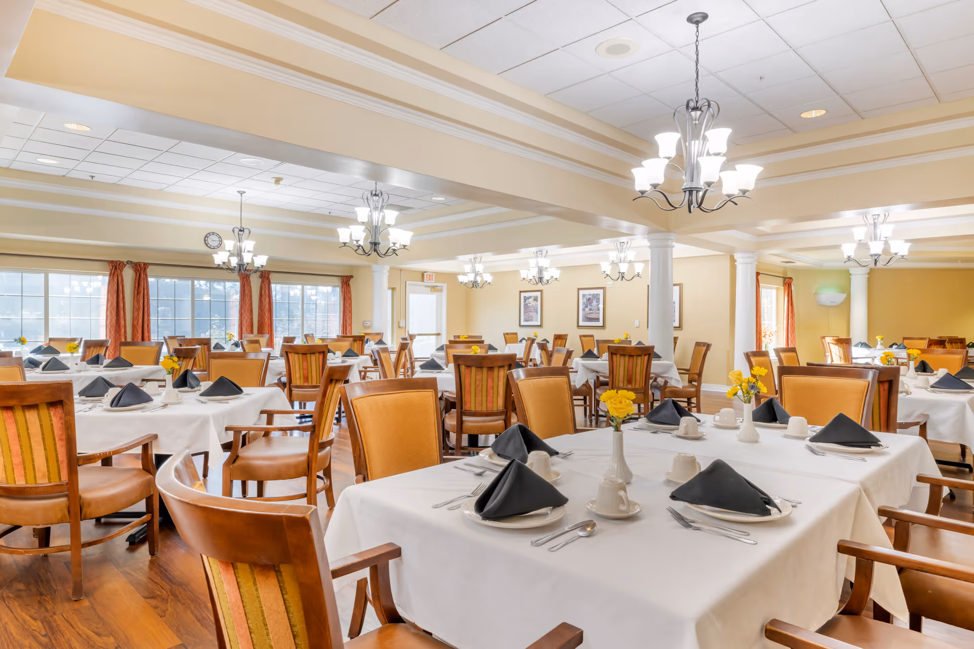 A spacious dining room with multiple tables covered in white tablecloths, each set with black folded napkins, white cups, plates, and silverware. The room features wooden chairs with striped upholstery, large windows with red curtains, chandeliers hanging from the ceiling, and yellow flowers in small vases on the tables.