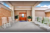 Covered outdoor seating area with white rocking chairs arranged along the sides, leading to a wooden double door entrance with glass panels. There are potted plants on either side of the door and a brick wall on the left side. The sky is partly cloudy.