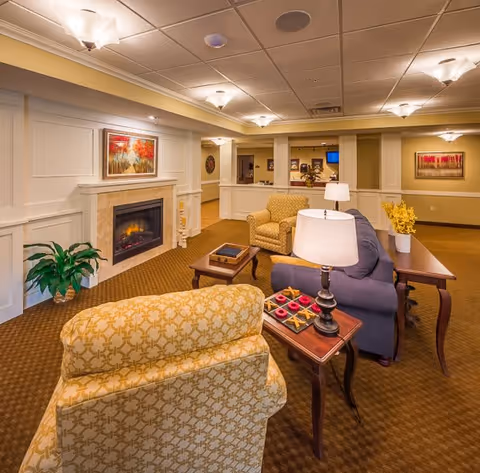A cozy living room area in a senior living facility with patterned armchairs, a blue sofa, wooden side tables with lamps, a fireplace with a painting above it, and warm lighting. The room has beige walls with white paneling and carpeted floors.