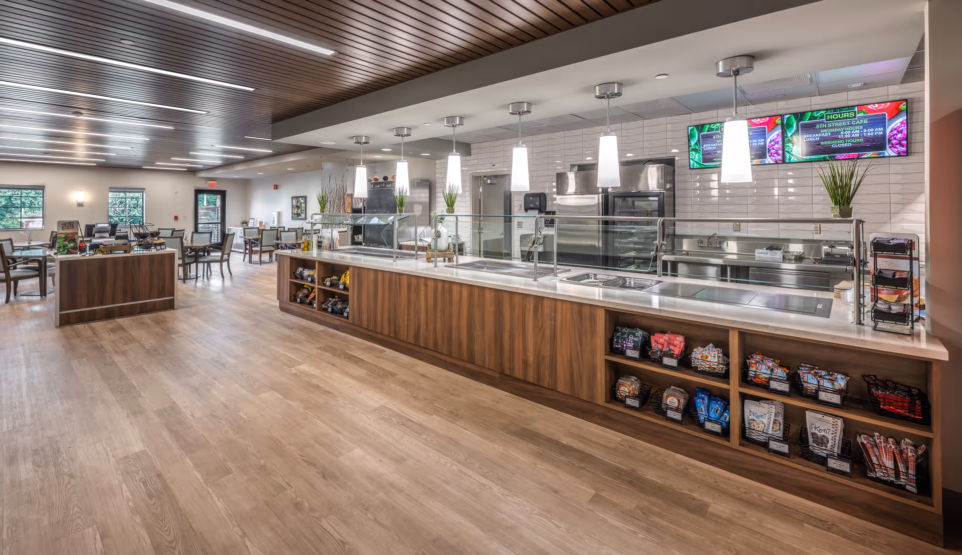 Spacious modern dining and serving area with a long wood-paneled service counter, glass sneeze guard, seating tables, and snack displays.