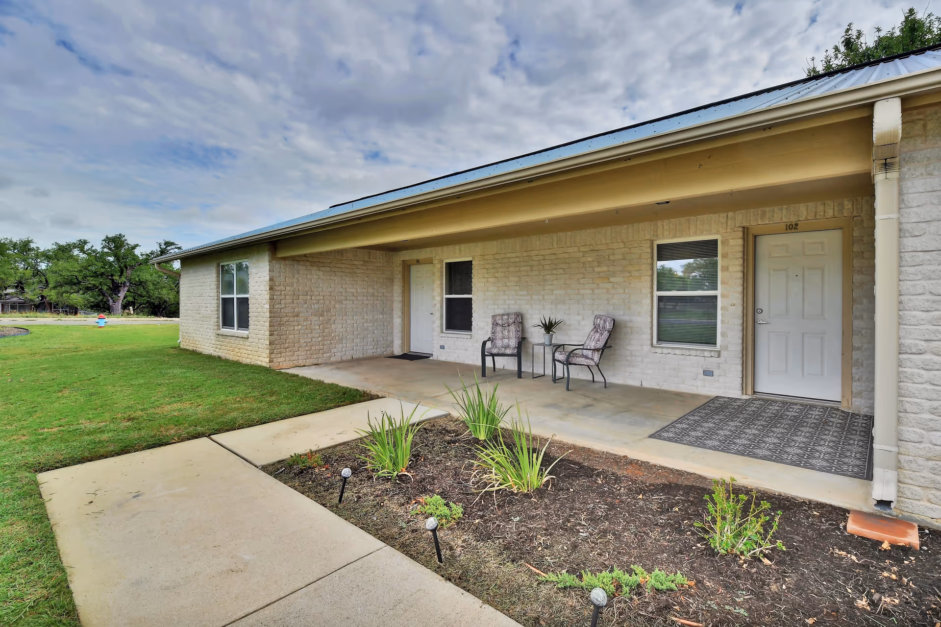 Front porch of a single-story brick unit with two chairs and a small table by a white door numbered 102, a concrete walkway and lawn.