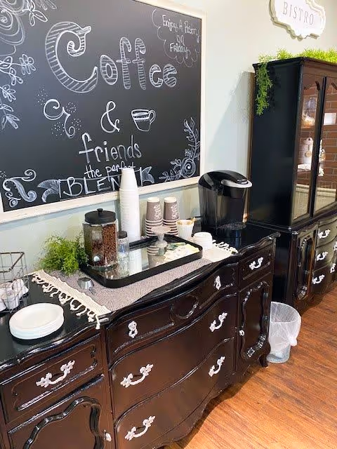 A coffee station setup with a black coffee maker, disposable cups, a container of coffee beans, and a small plant on a dark wooden cabinet. Above the cabinet is a chalkboard sign with decorative writing that says 'Coffee & friends the perfect blend' and a small sign that reads 'Bistro'.