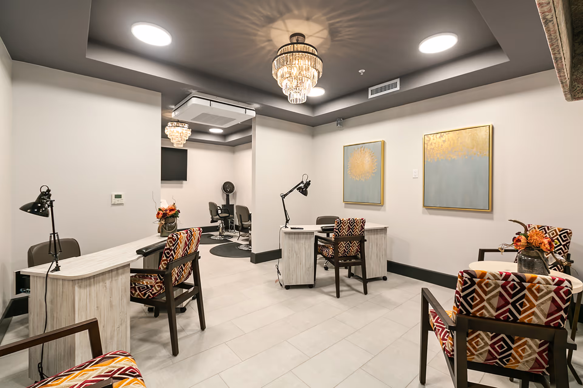 Interior view of a modern salon area in a senior living facility with patterned chairs, two desks with lamps, a small seating area with flowers, and two framed abstract paintings on the wall. The ceiling has recessed lighting and decorative chandeliers.