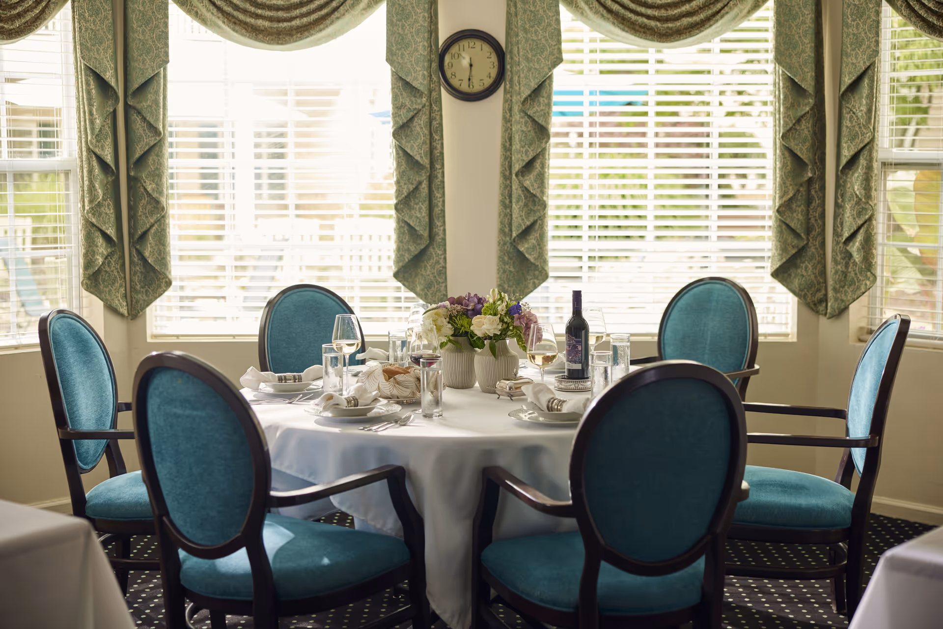 A round dining table set for six with teal upholstered chairs in a room with large windows covered by green patterned curtains. The table is covered with a white tablecloth and set with plates, glasses, napkins, and a centerpiece of flowers. A bottle of wine and wine glasses are also on the table. A clock is mounted on the wall between the windows.